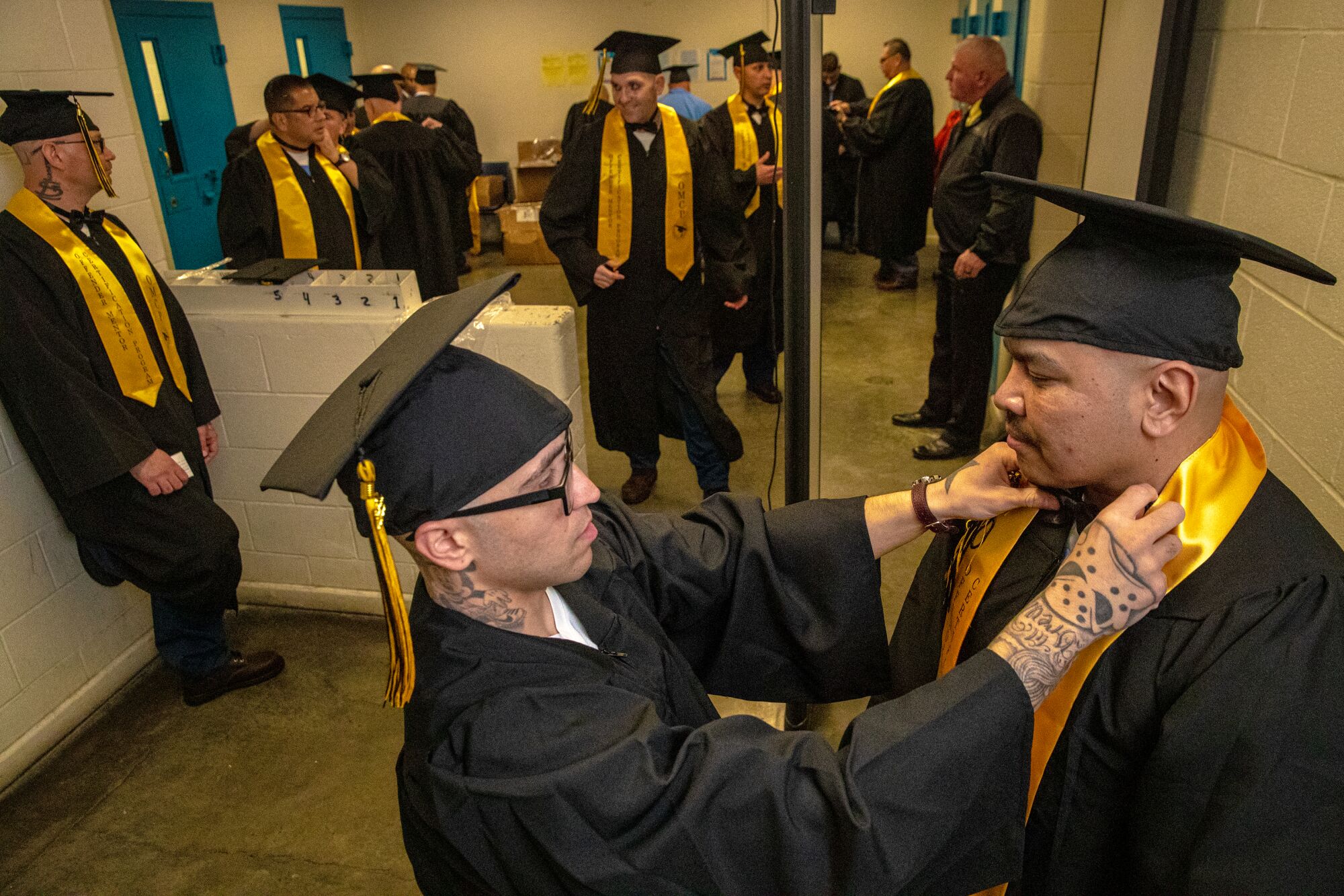 A man in cap and gown adjusts the gold sash around the neck of another man in cap and gown 