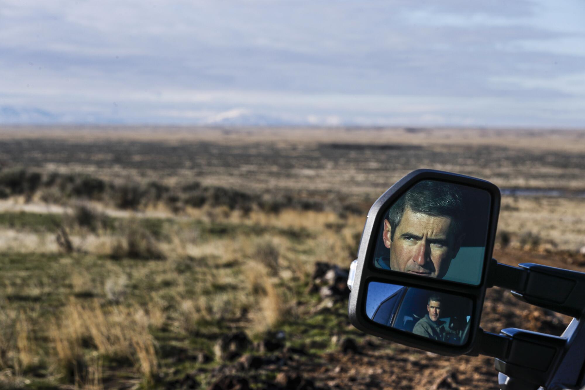 A man's face is reflected in a side-view mirror as he drives across a proposed wind farm site.