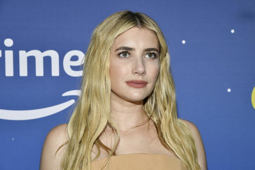 Emma Roberts gazes upwards while wearing a strapless dress while standing in front of a blue background