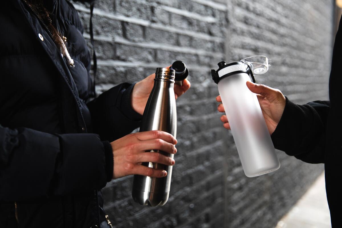 Two women stand next to a black wall, holding reusable water bottles.