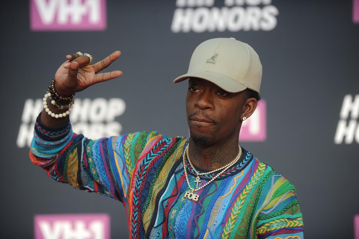 Rich Homie Quan in a muted green hat and a colorful sweater holding up a peace sign and posing in front of a black background