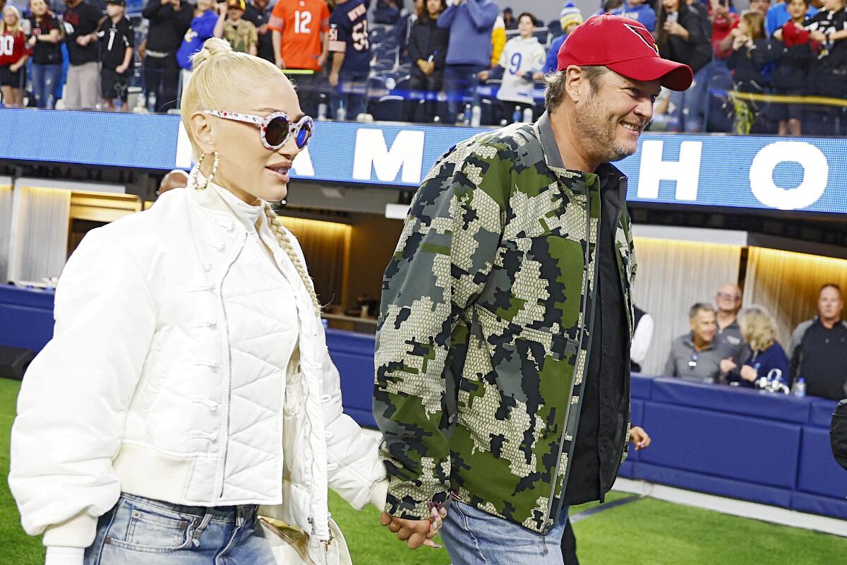 Gwen Stefani, in a white jacket and sunglasses, holds hands while walking with a camo-jacketed Blake Shelton