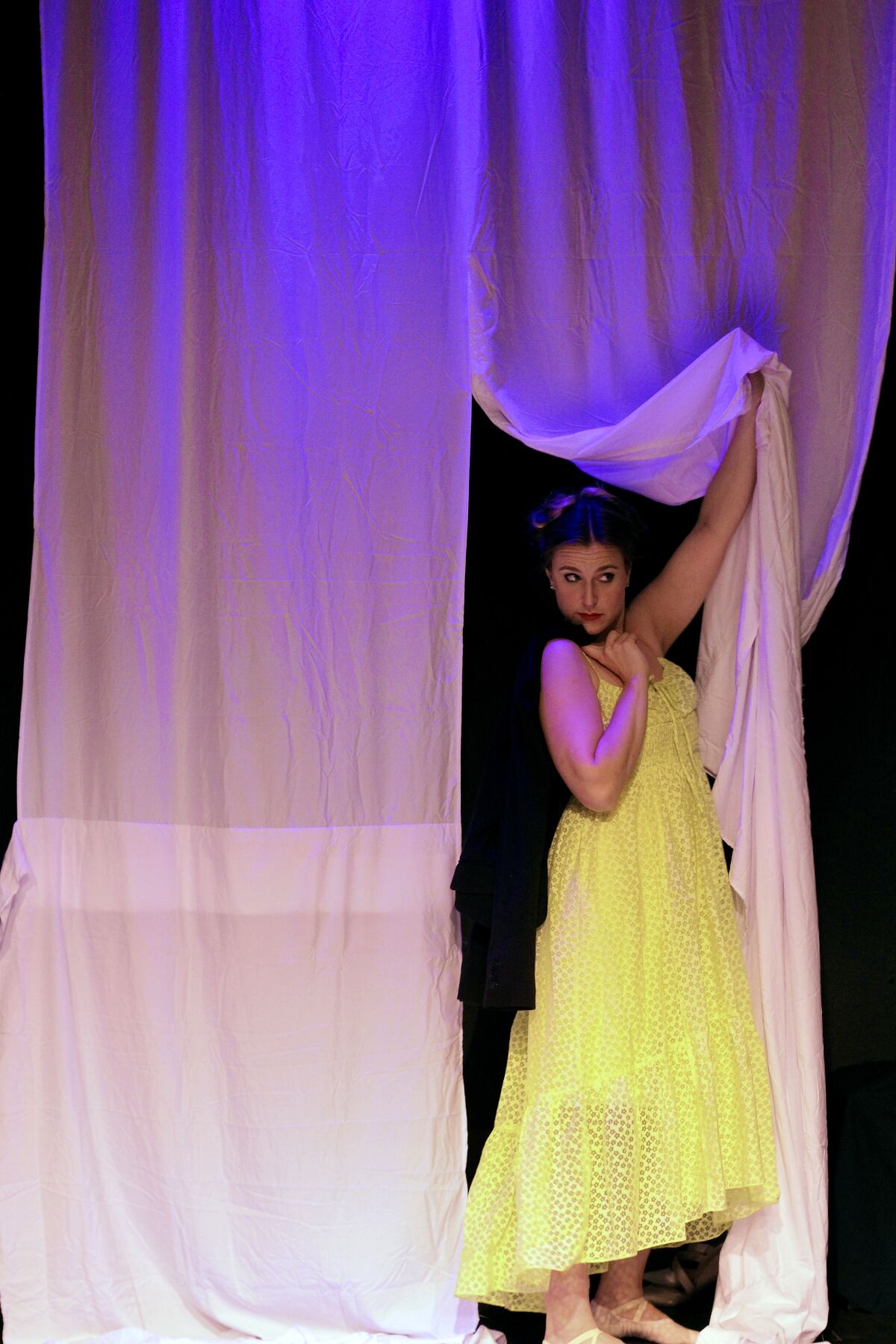 A woman in a yellow gown peeks around a curtain.