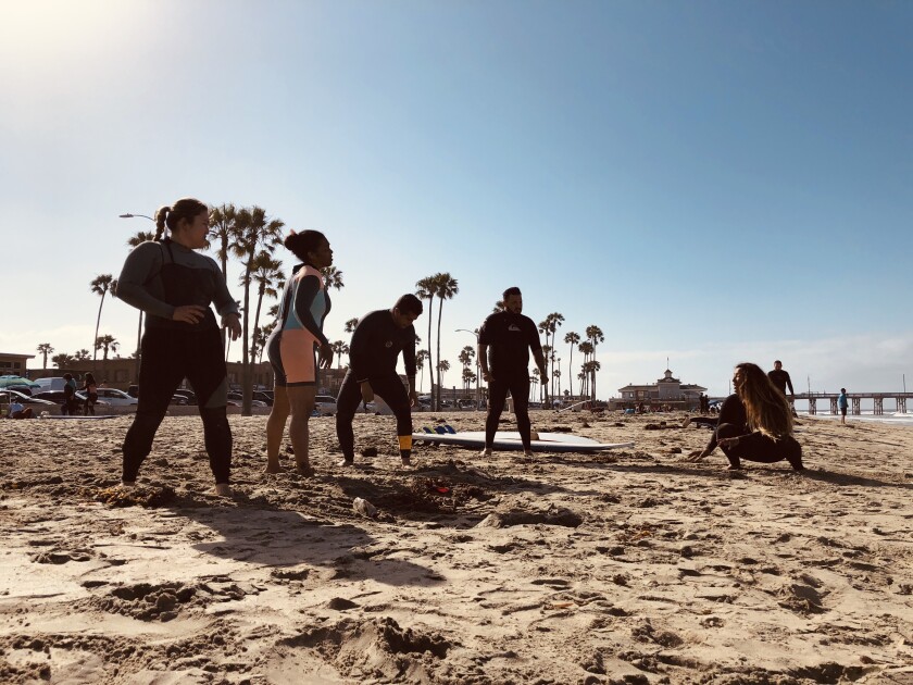 Four people stand on a sandy beach as another person crouches in front of them.