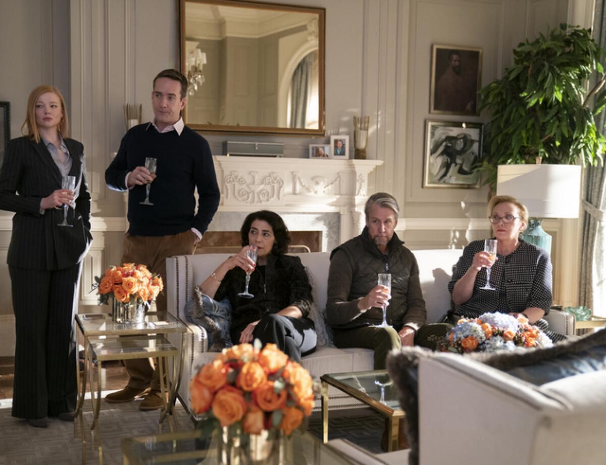 A man and a woman stand with drinks in their hands while three people sit on a couch in a scene from "Succession."