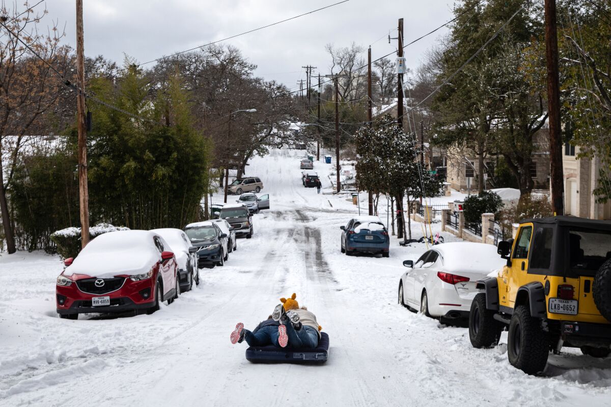 Two people use an air mattress to slide down a snow-covered street.