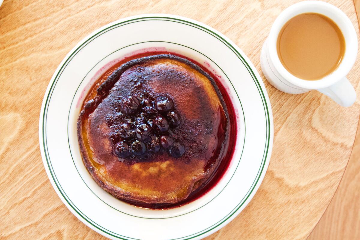 An overhead photo of brown-butter pancakes with blueberry compote on a wooden table with a mug of coffee