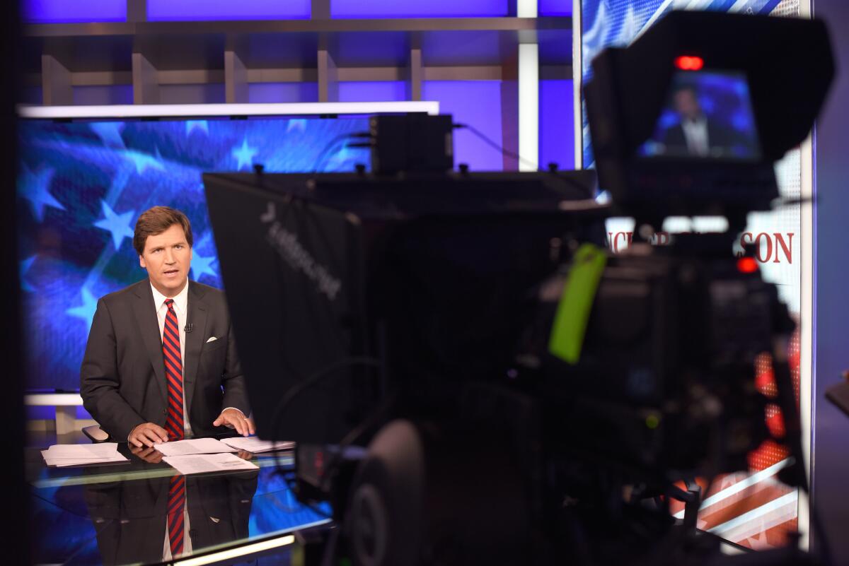 Fox News' Tucker Carlson sits at a news desk with a camera in the foreground