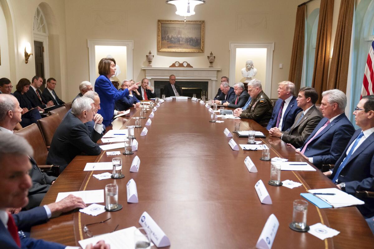 Nancy Pelosi stands at a table full of men and speaks to Donald Trump.