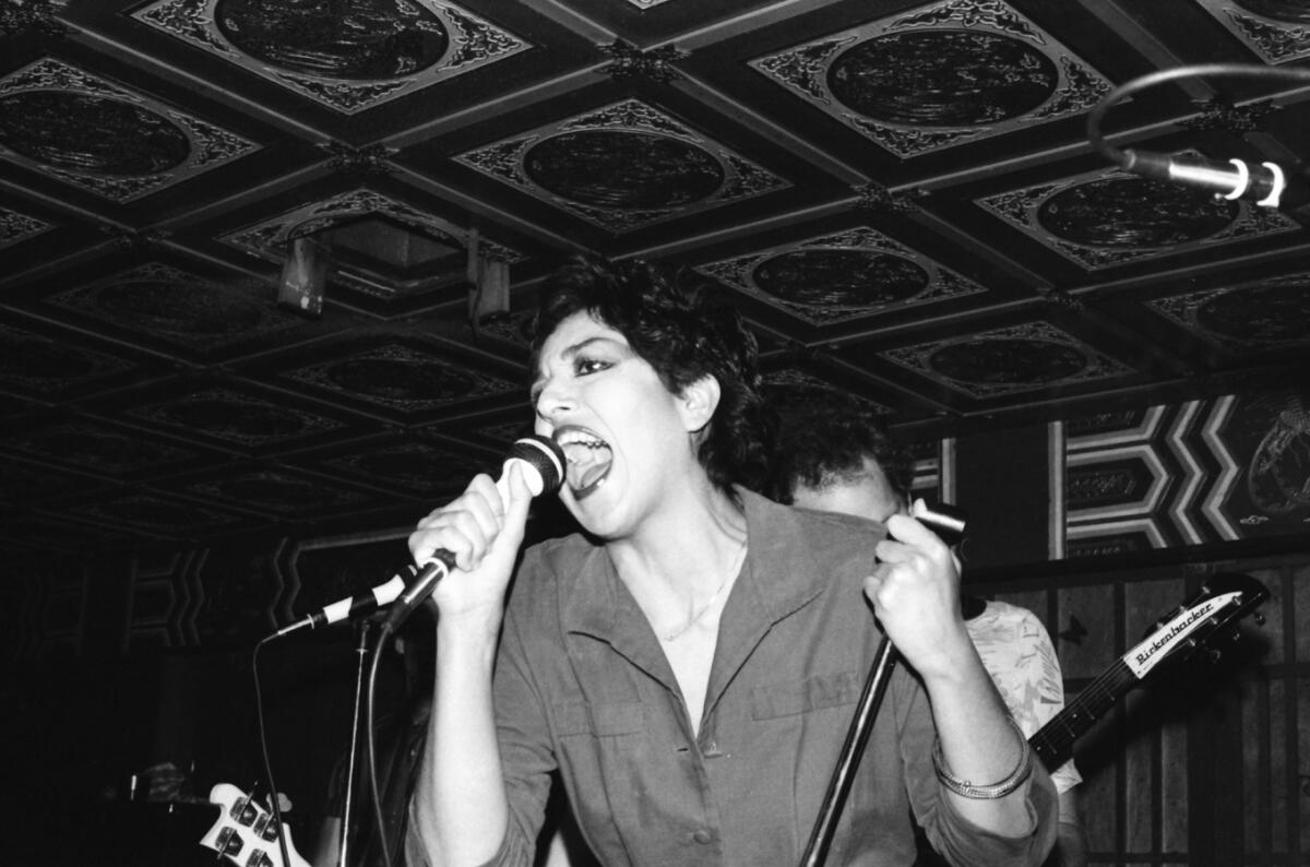 Alice Bag holds a microphone to her mouth and the mic stand with her left hand.
