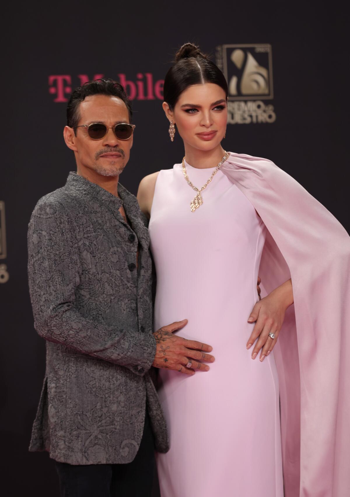 Marc Anthony in a gray suit posing with his hand on wife Nadia Ferreira's belly. She's wearing a light pink dress.