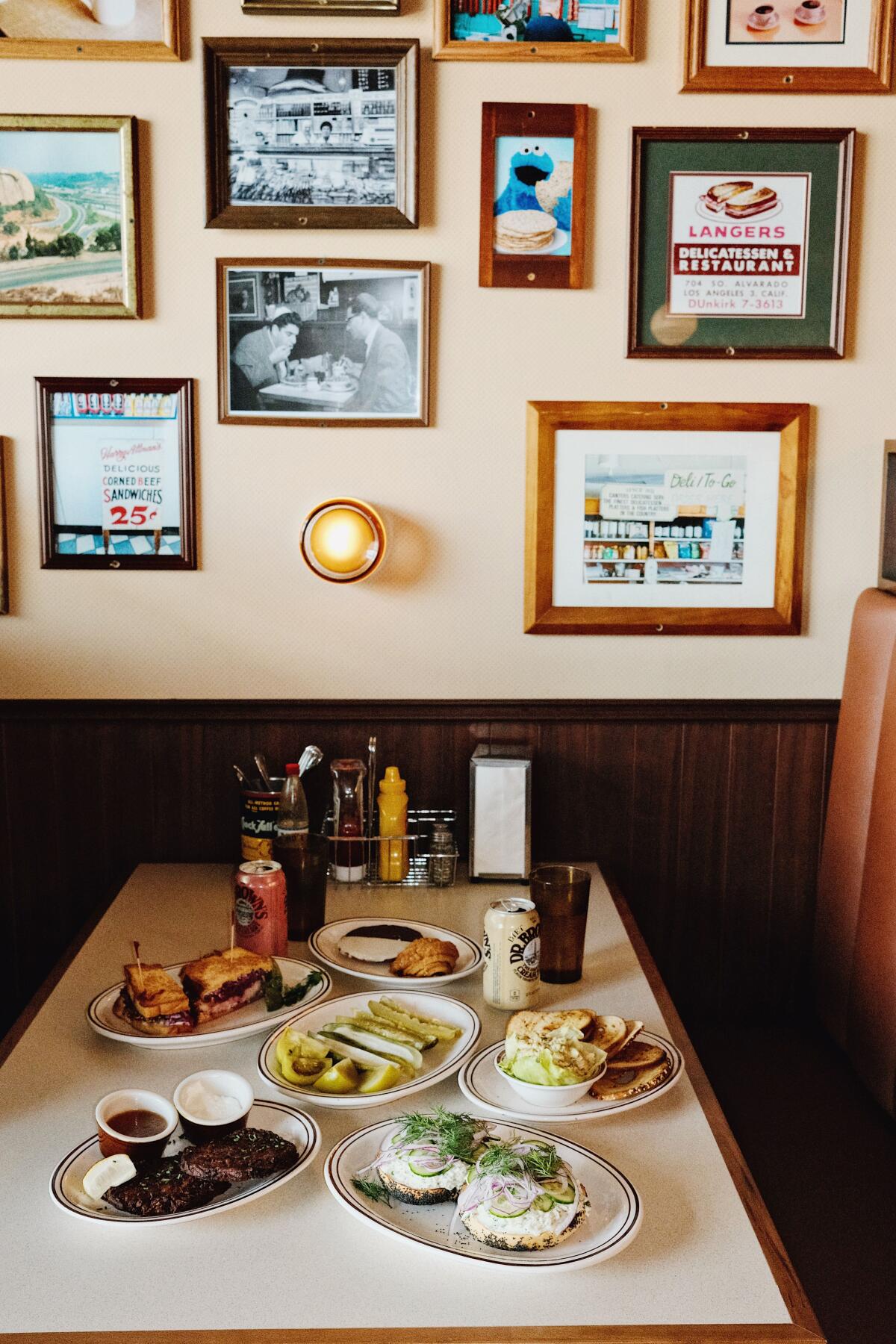 A booth full of dishes from Belle's Bagels Delicatessen and Bar: sandwiches, pickles, cookies and more. Pictures on wall