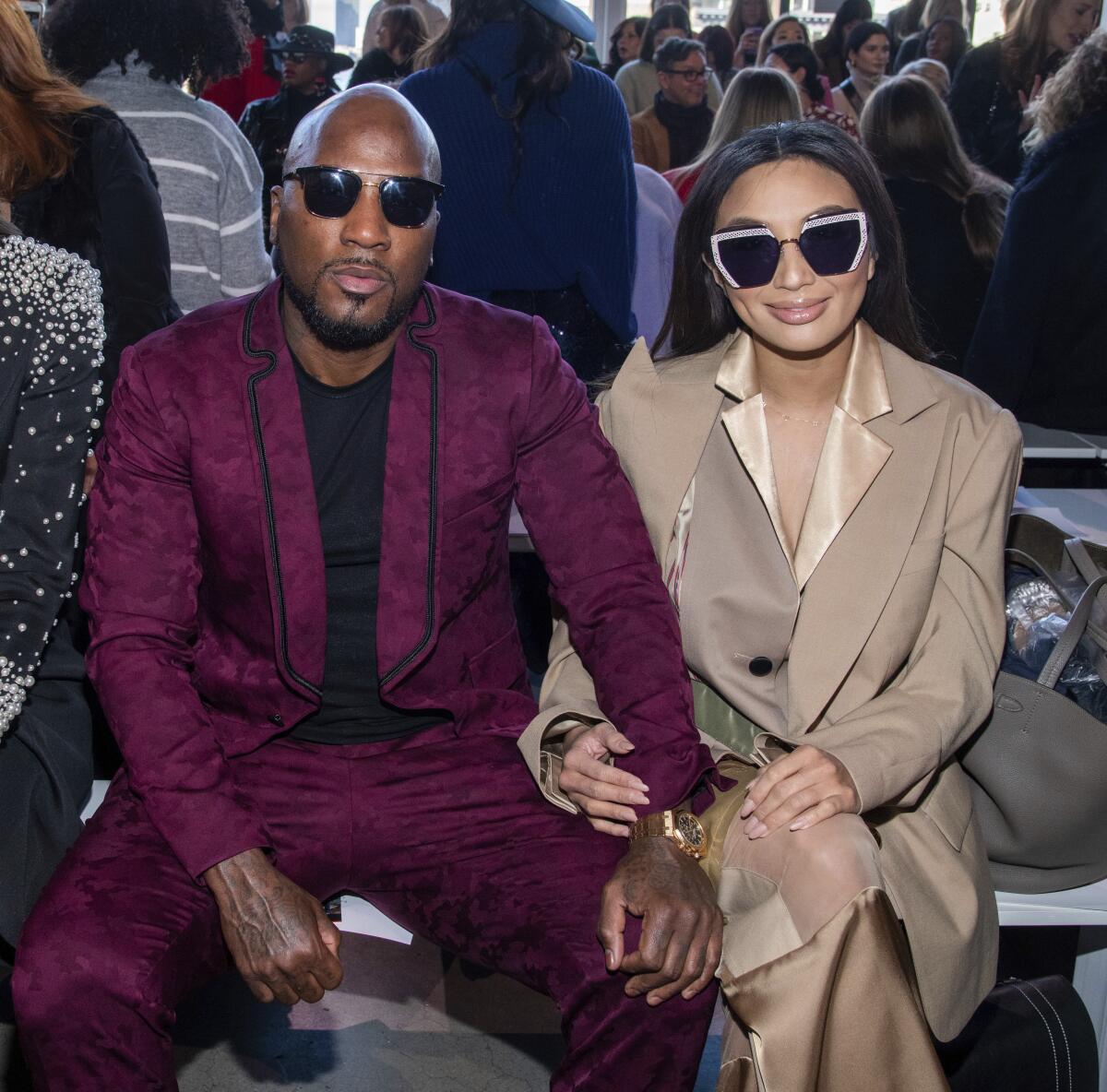 A bald Black man in a maroon suit and sunglasses sits with an Asian woman with long hair in a beige suit and sunglasses.