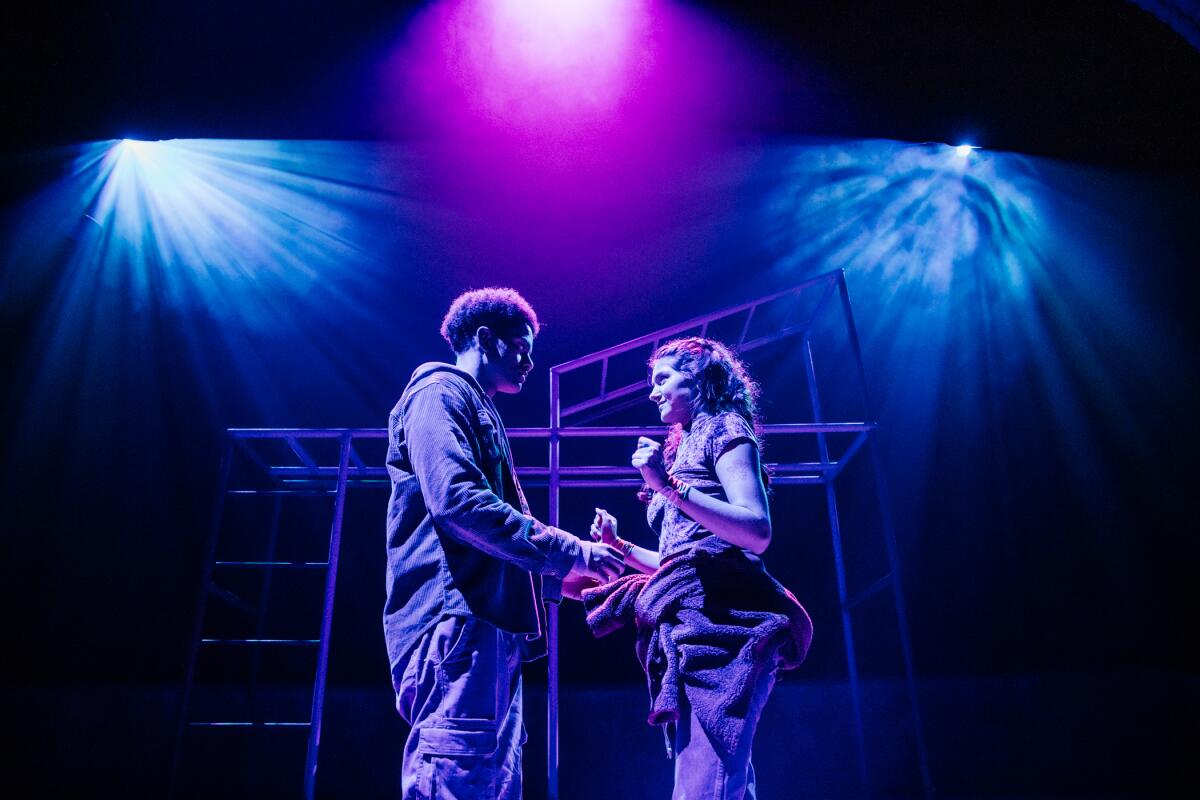 A young man and woman face each other on a stage under blue and purple lights.