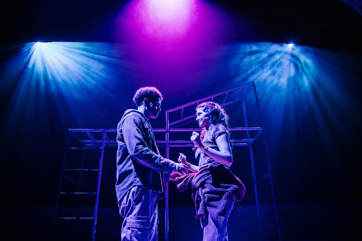 Two actors dressed in casual outfits stand on a sparsely furnished stage bathed in blue and lavender light.