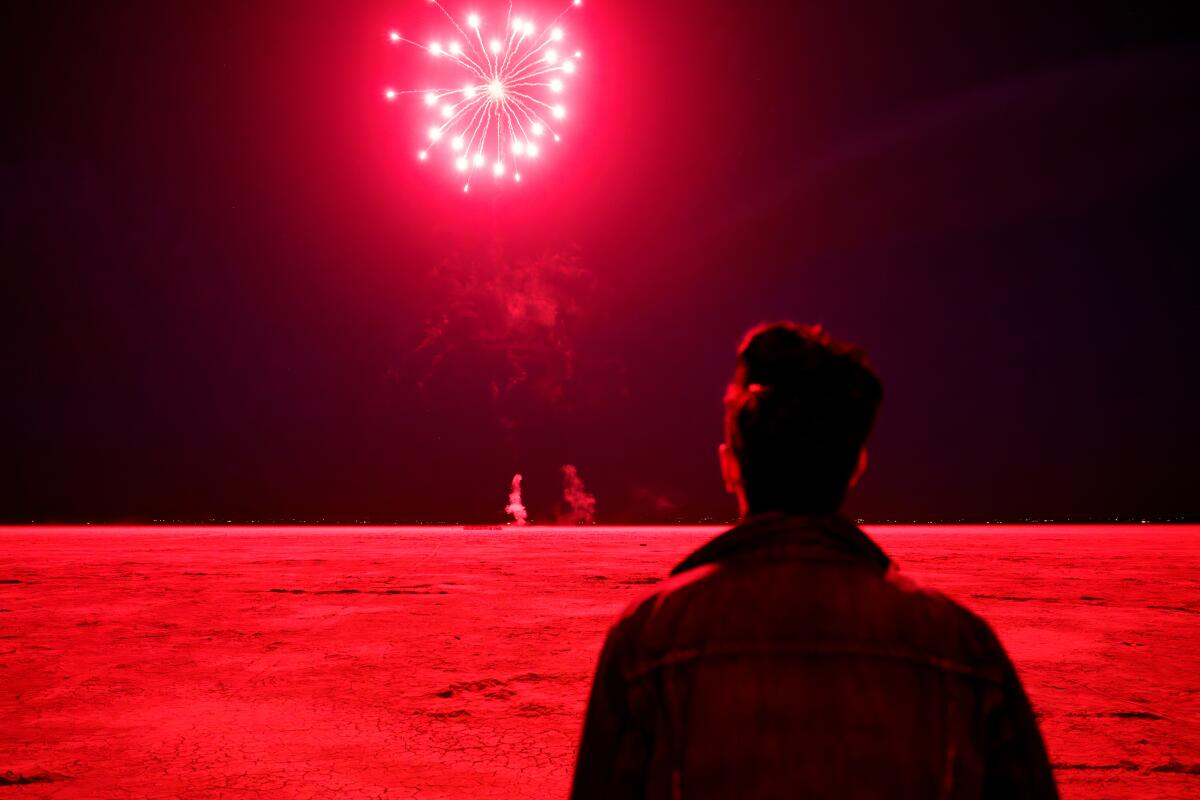A man's back is turned to the camera as he looks out toward a red firework.
