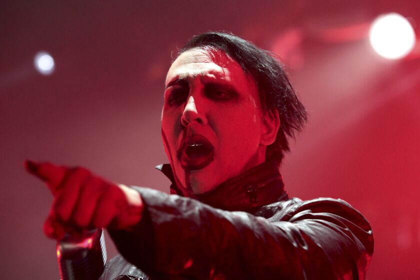 A man in Goth makeup and a leather jacket points forward while performing on a stage filled with red light