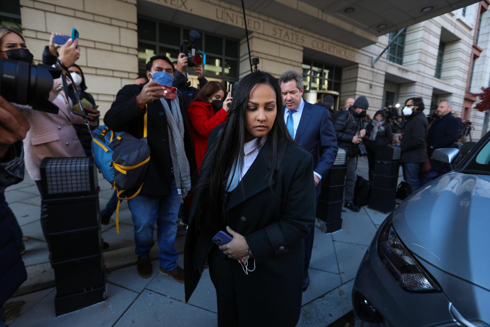  A woman with long dark hard stands near the front of a vehicle outside a courthouse as people hold up phones and cameras.  