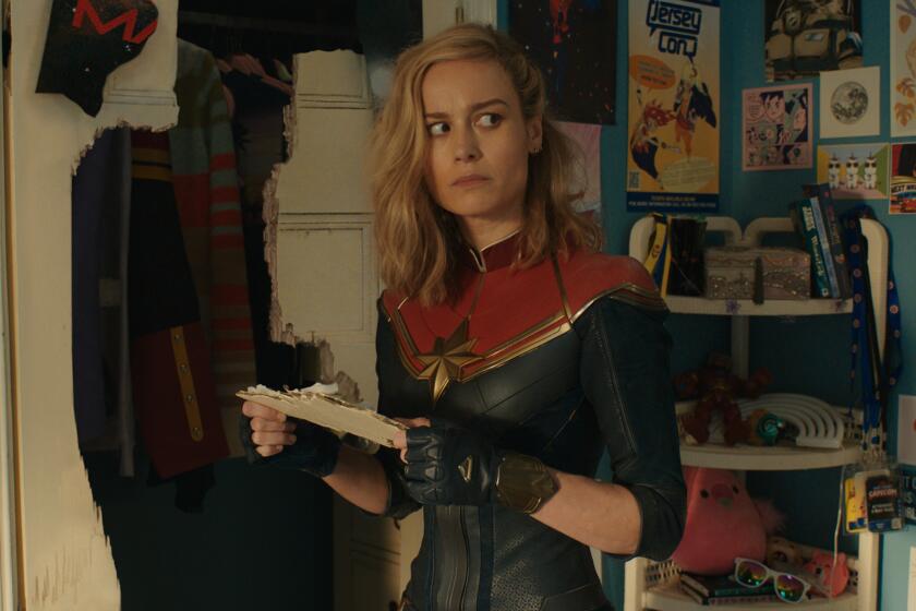 Brie Larson holds a piece of paper in a room covered in posters. She wears a Captain Marvel costume
