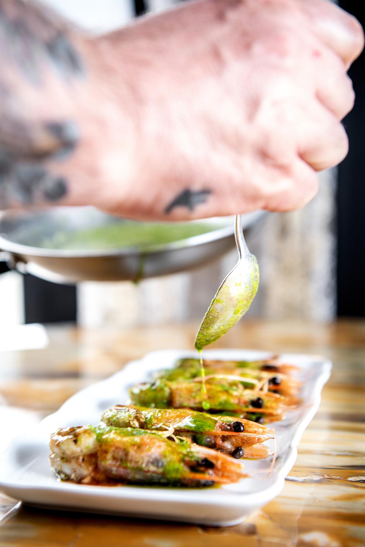 A hand holding a spoon drizzles salsa verde onto prawns.