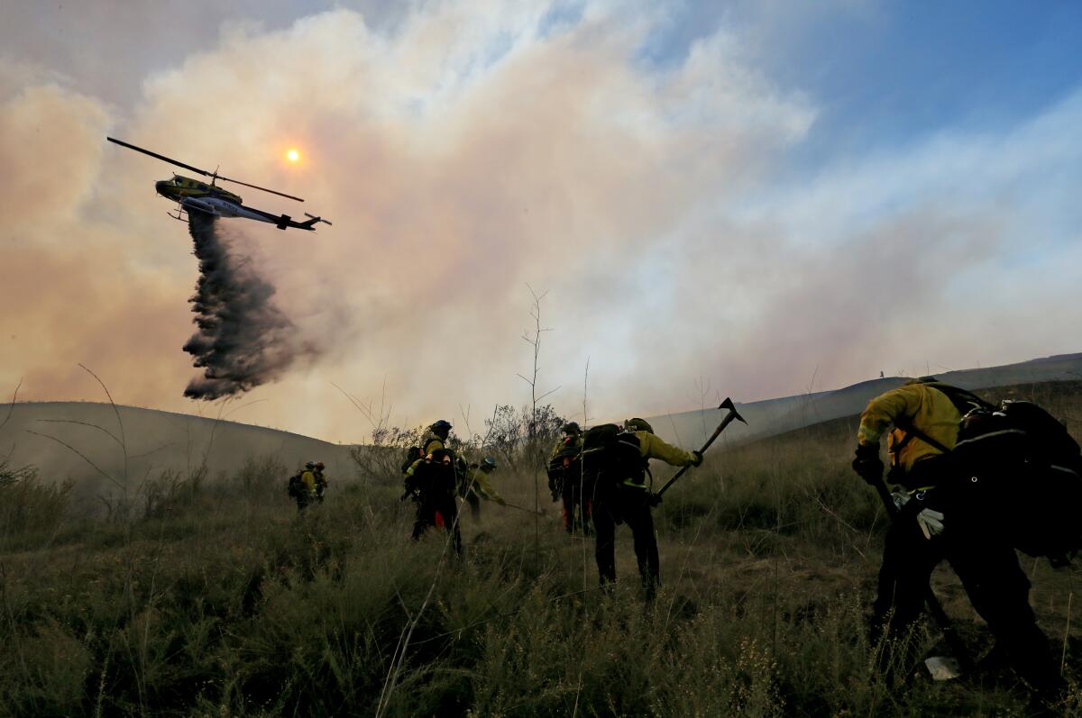 Smoke dims the sun as a helicopter drops water. Firefighters use hand tools to cut a fire break on a hillside.