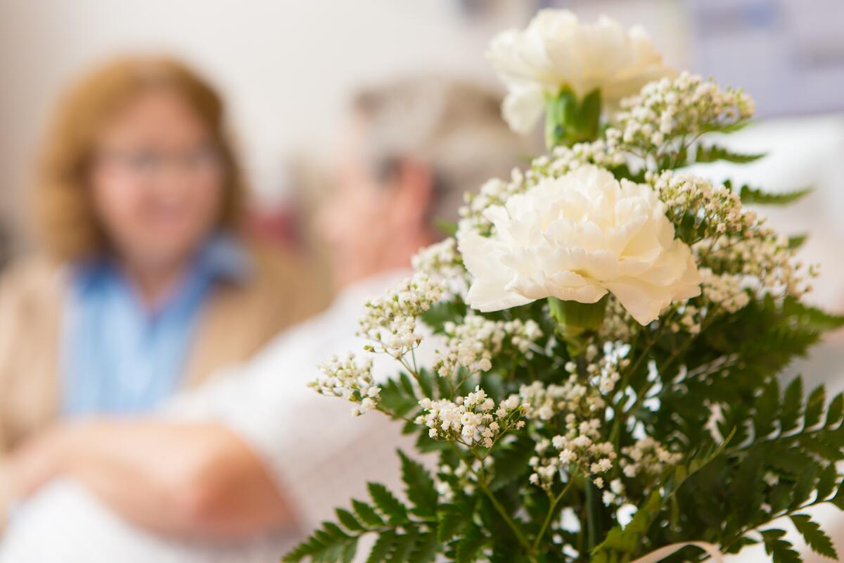 A woman visits a patient in the hospital, with a bouquet of flowers in the foreground.