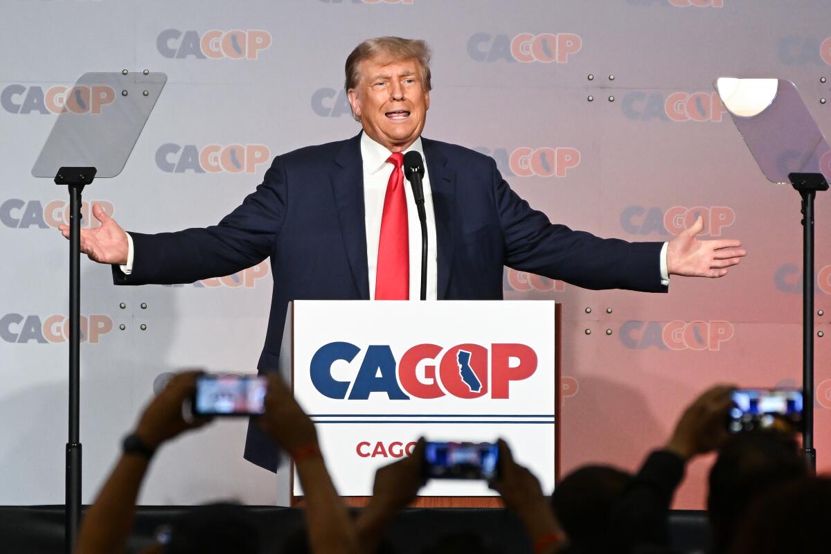 A man in a dark suit and red tie holds out his arms at a lectern with the words CAGOP 
