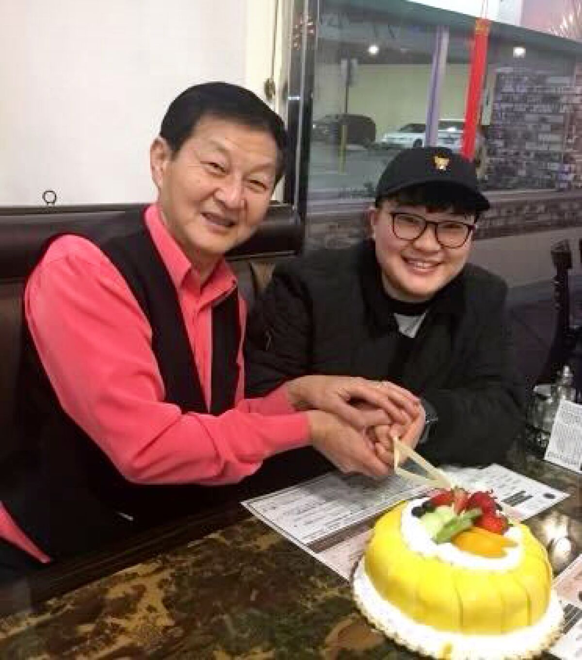 Two people are seated, smiling looking at camera, both holding the same knife, about to cut a cake with fruit on top.