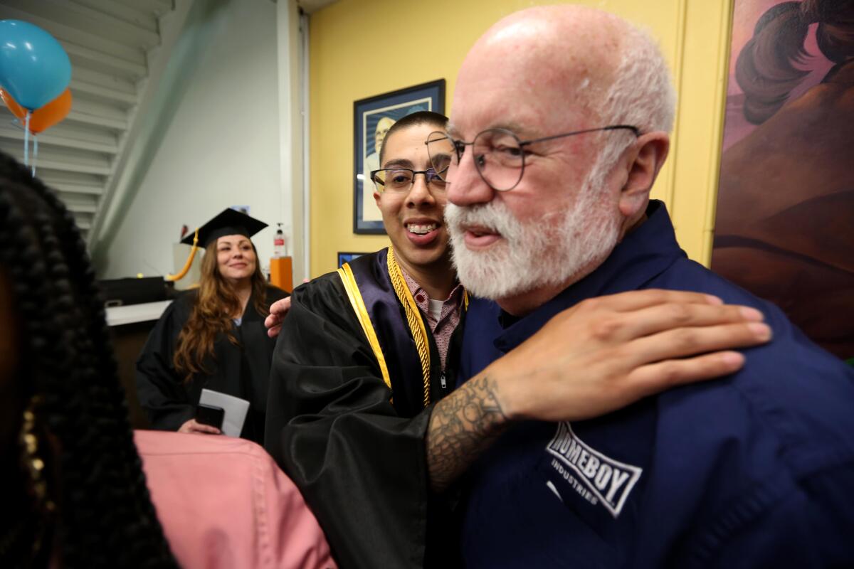 A younger man in graduation attire hugs an older man with a white beard wearing a shirt with the Homeboy logo.