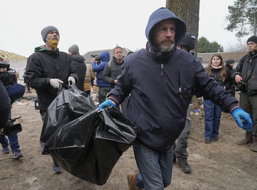 A man, on the right, wearing a hooded jacket and blue gloves, carries a black body bag with the help of another man.