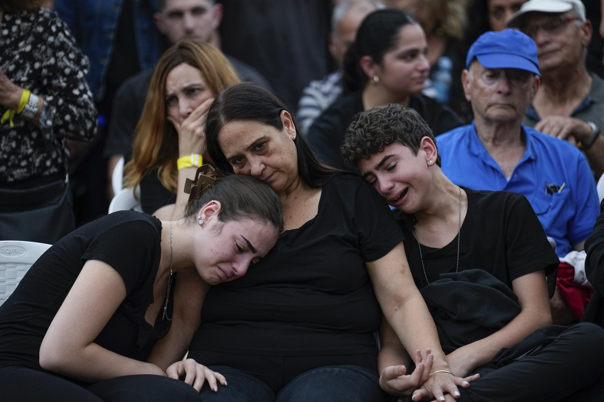 A woman wearing black is flanked by a young woman, left and a young man, both wearing black and crying, near other people