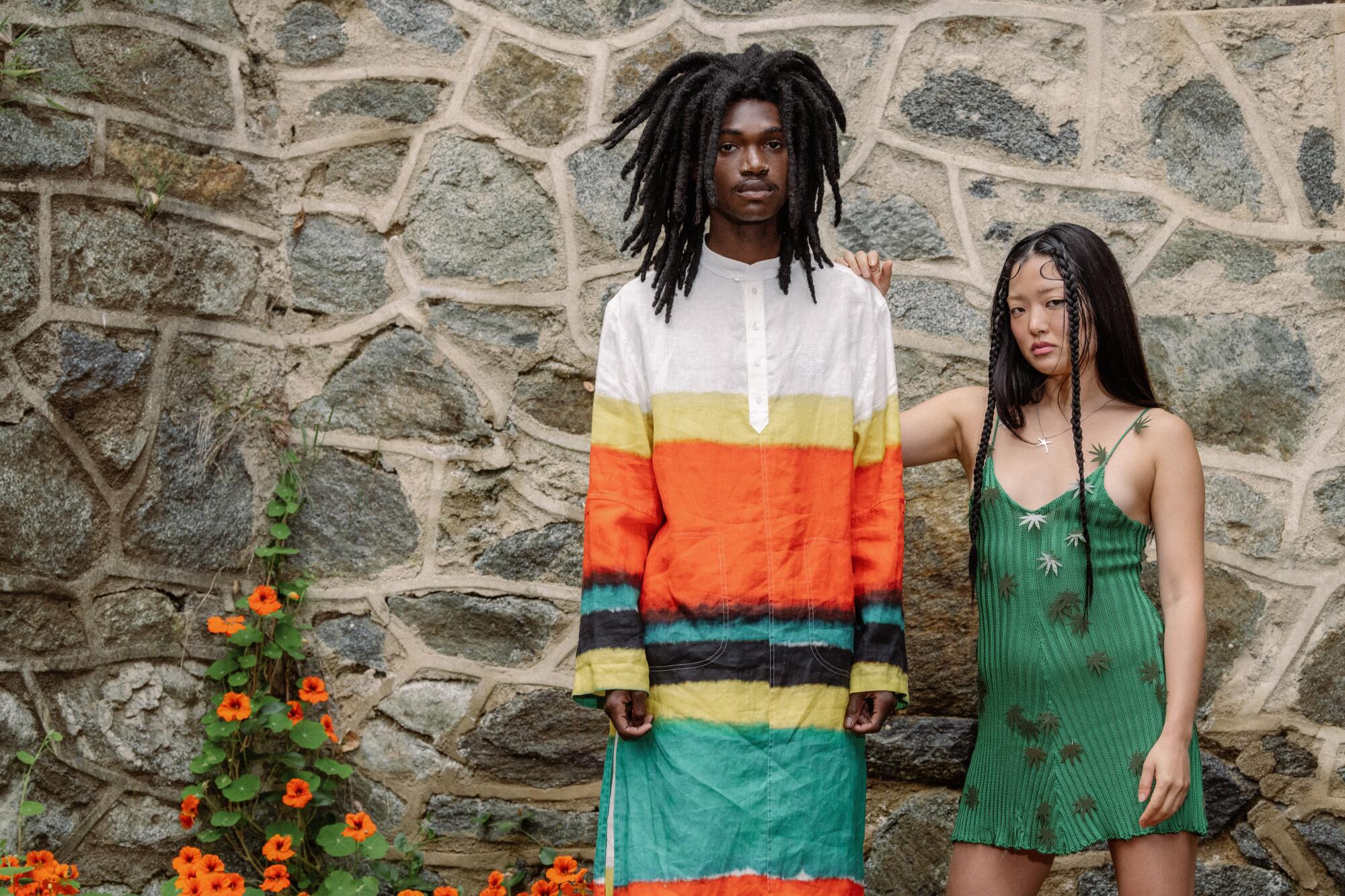 man in colorful striped outfit, left and woman in green dress pose outside 