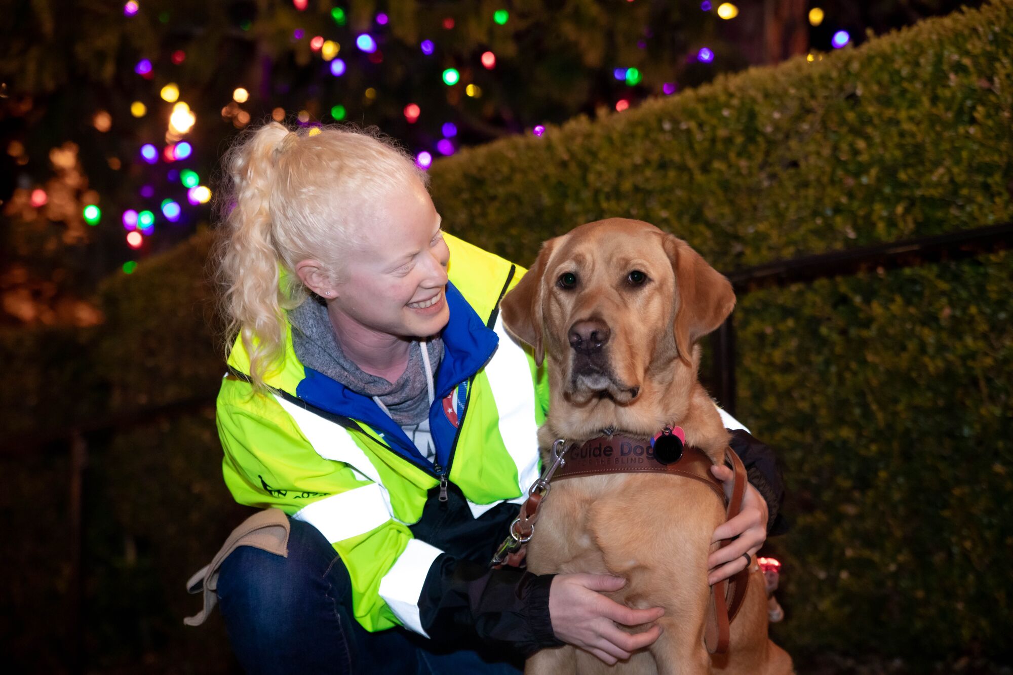 Kym Crosby and guide dog Tron pose in front of holiday lights.