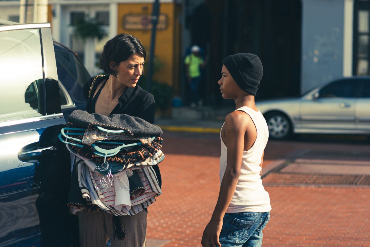 A woman standing by an SUV holds an armload of clothing on hangers while talking to a young boy.