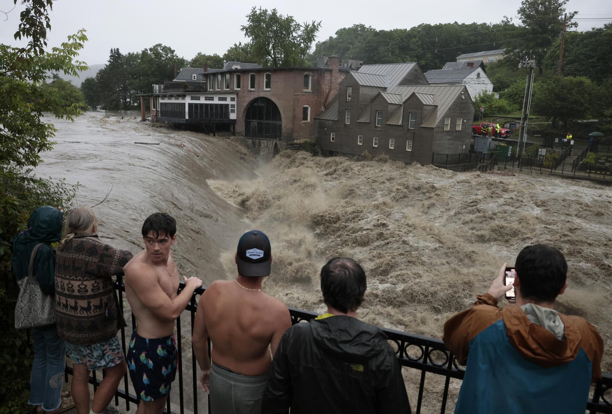 Several people standing at a railing overlooking an expanse of muddy, roiling water as it passes historic buildings.