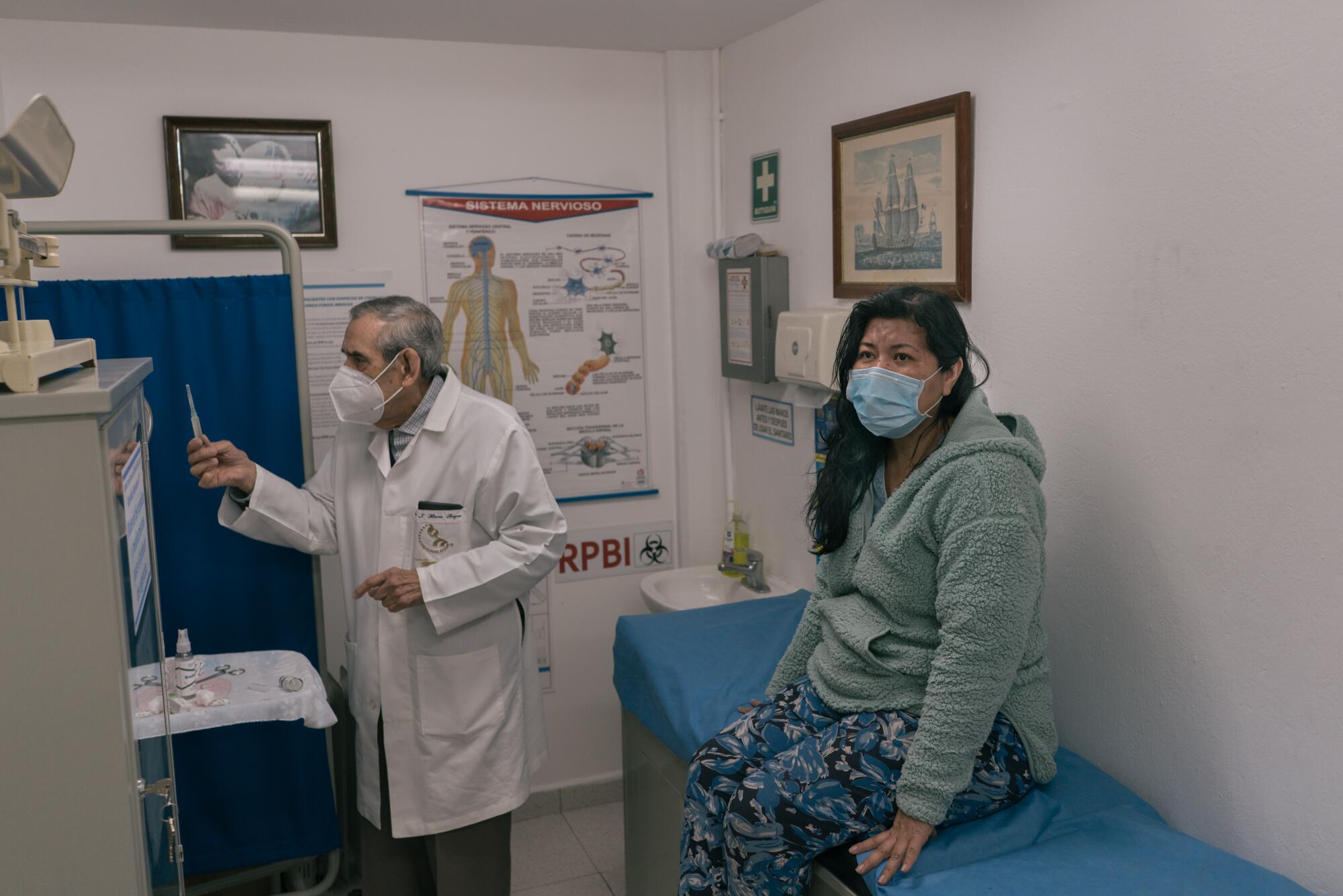 A masked man in a white coat holds a syringe near a masked woman seated on an examination table 