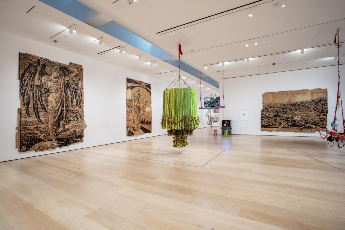 A museum gallery with three works on cardboard on the walls and a hanging green chandelier in the center