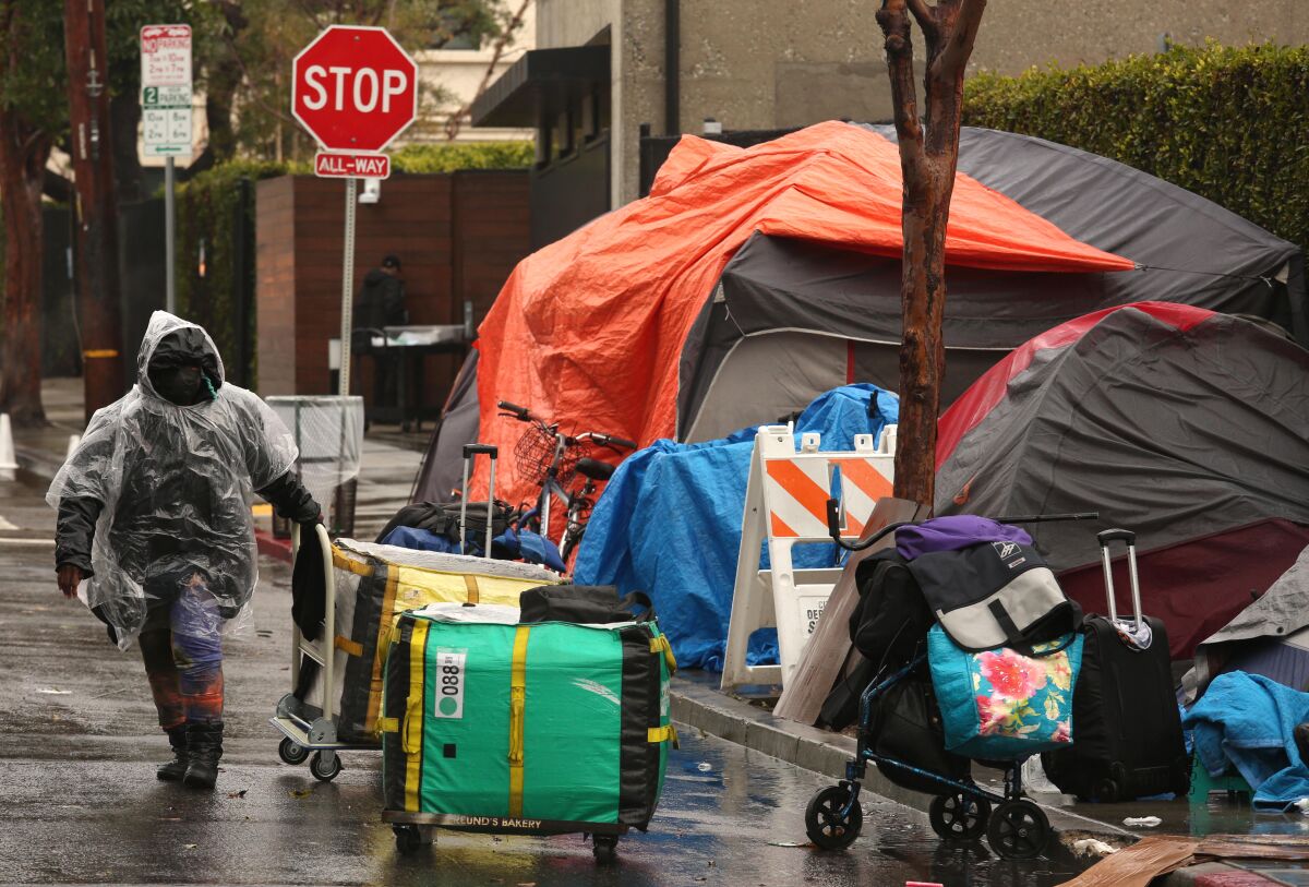 A person in a plastic raincoat leads his belongings along a wet street past a group of tents on the sidewalk.
