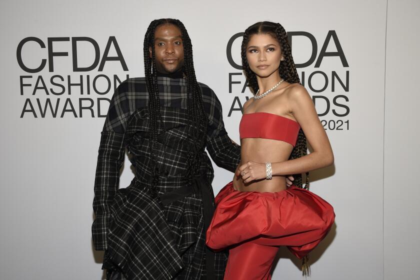 A man with braids in a dark plaid gown and Zendaya in a bright red bandeau and puffy skirt