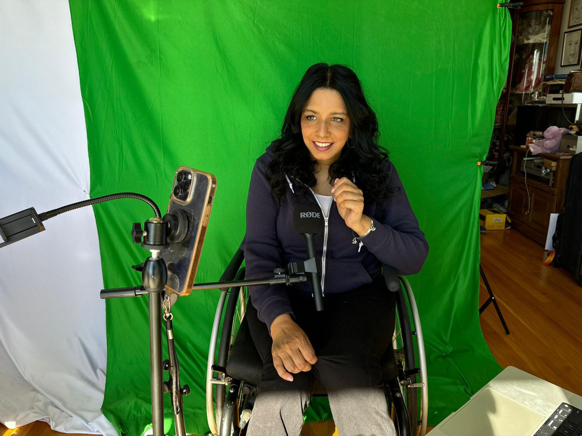 A woman dressed in dark clothes, sitting in a wheelchair against a green background, films herself with her mobile phone.