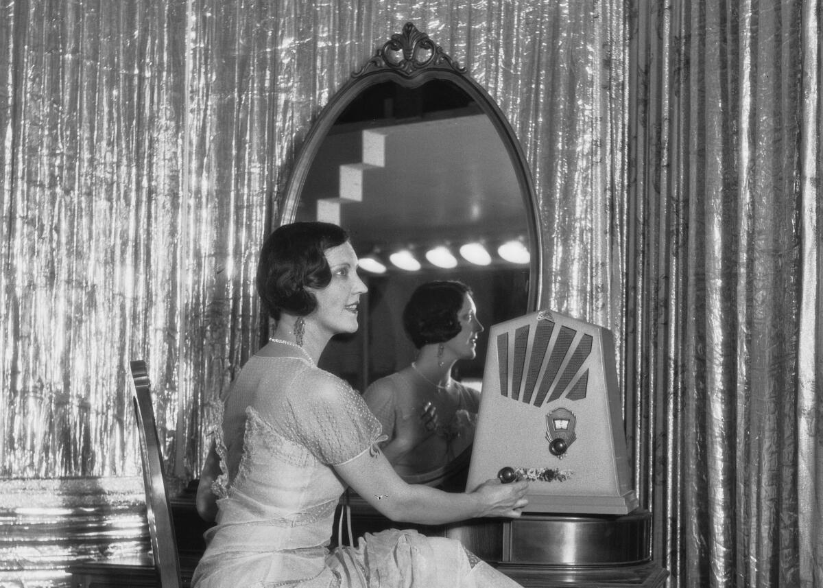 A woman in a frilly dress sits before a large vanity mirror with an ornate radio.