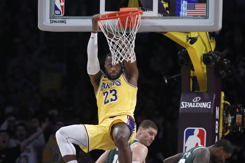 LOS ANGELES, CALIF. -- FRIDAY, MARCH 6, 2020: Los Angeles Lakers forward LeBron James (23) hangs from the rim after slam dunking the ball guarded by Milwaukee Bucks forward Marvin Williams (20) and Milwaukee Bucks center Brook Lopez (11) in the first half at the Staples Center in Los Angeles, Calif., on March 6, 2020. (Gary Coronado / Los Angeles Times)