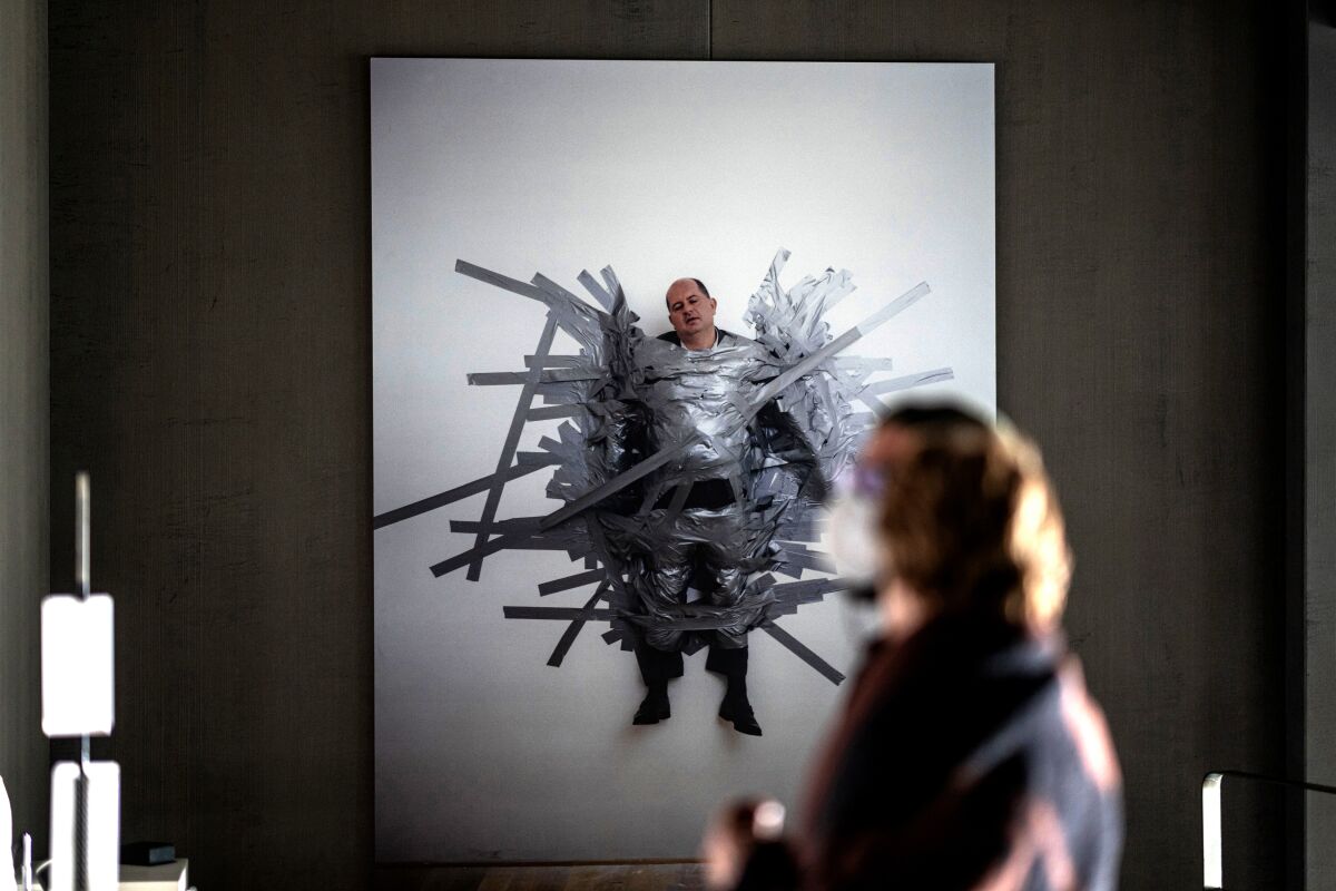 A person stands before an artwork that features a man duct-taped to a white canvas.