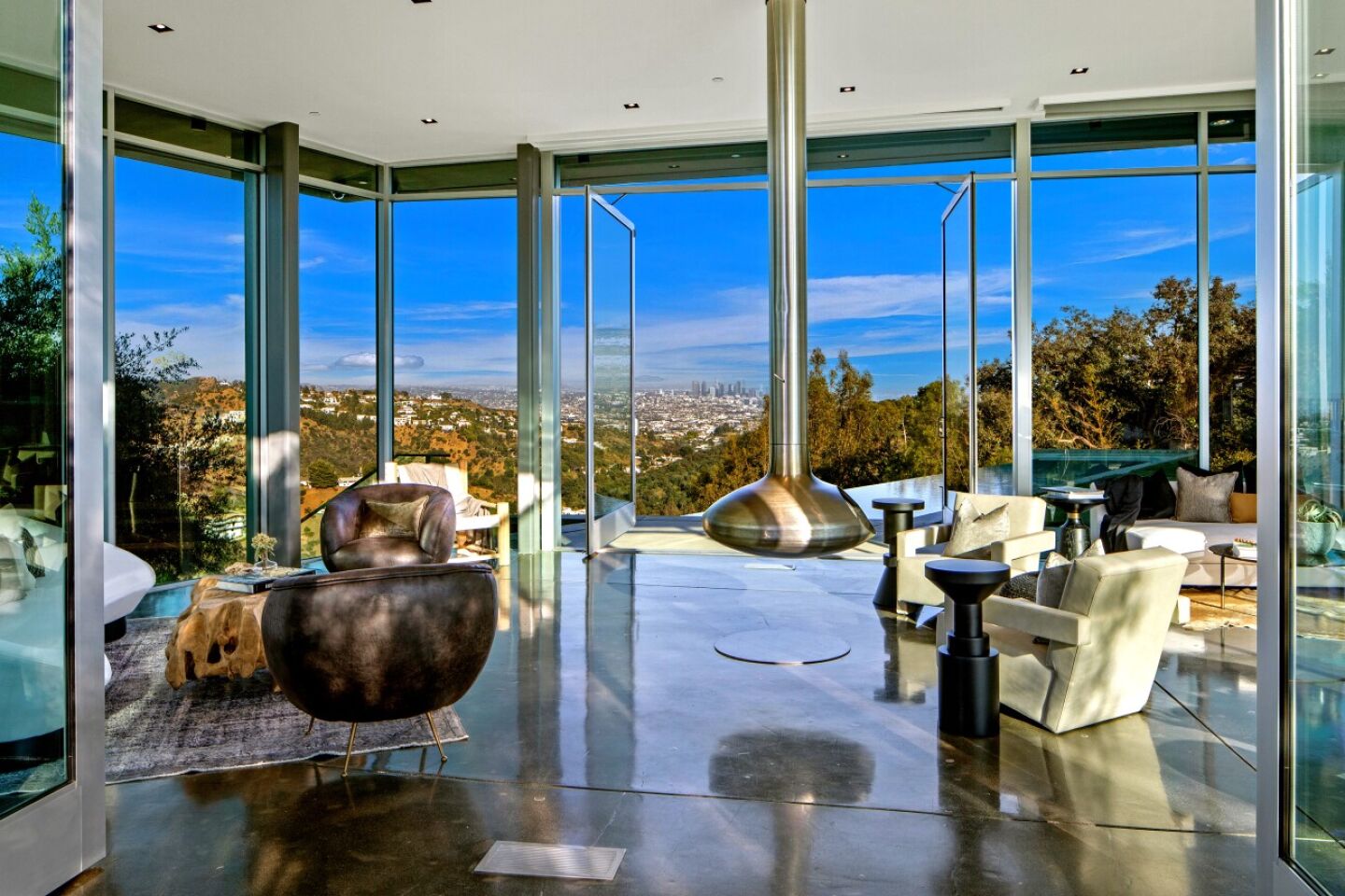 The living room has glass walls, a shiny floor and furniture overlooking greenery and sky.