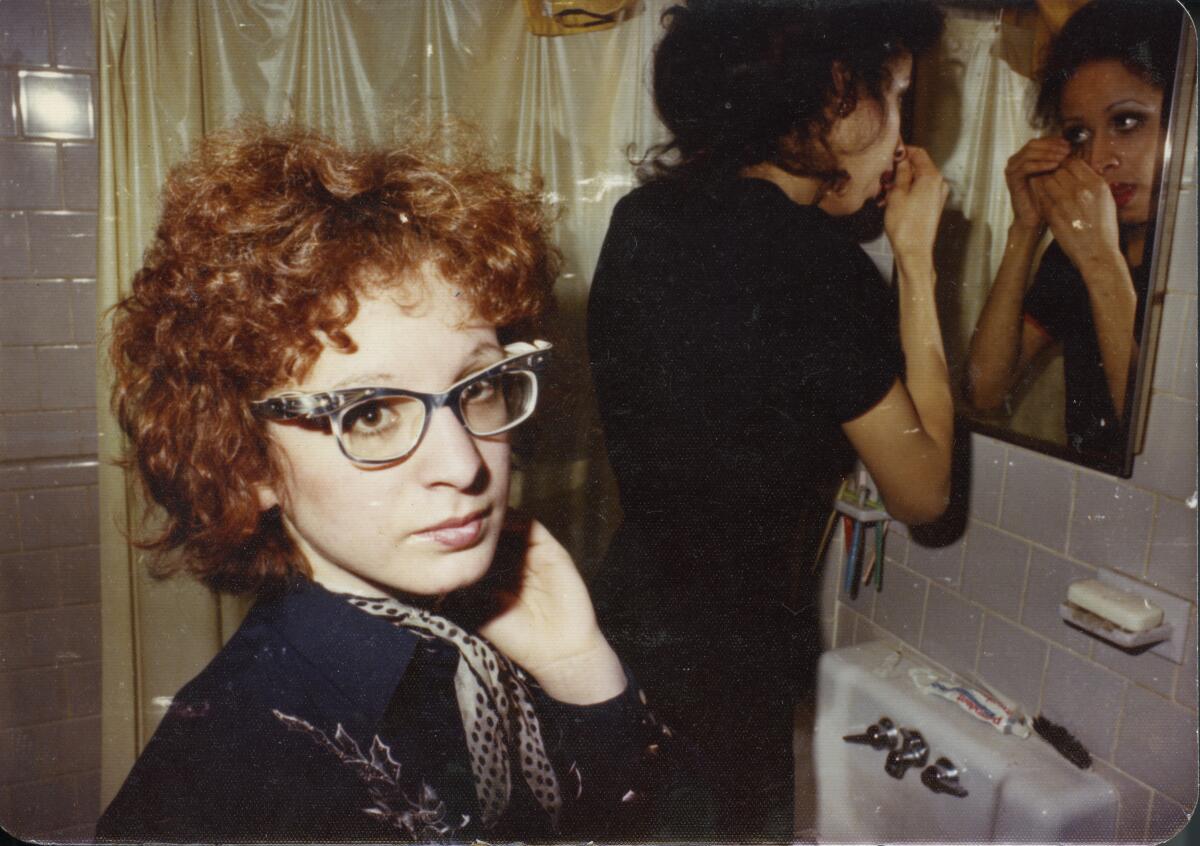 A red haired woman in glasses looks at the camera and another looks at a mirror in "All the Beauty and the Bloodshed."