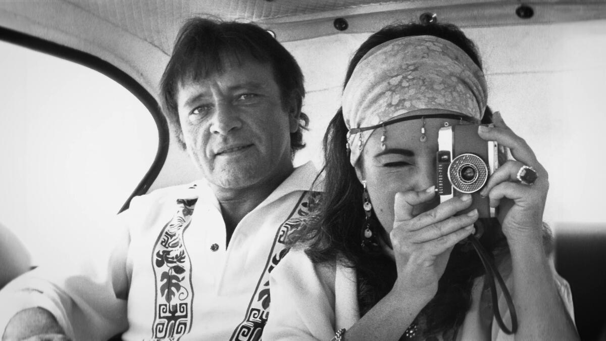 Richard Burton sits next to Elizabeth Taylor in a car as she holds a camera in front of her face.