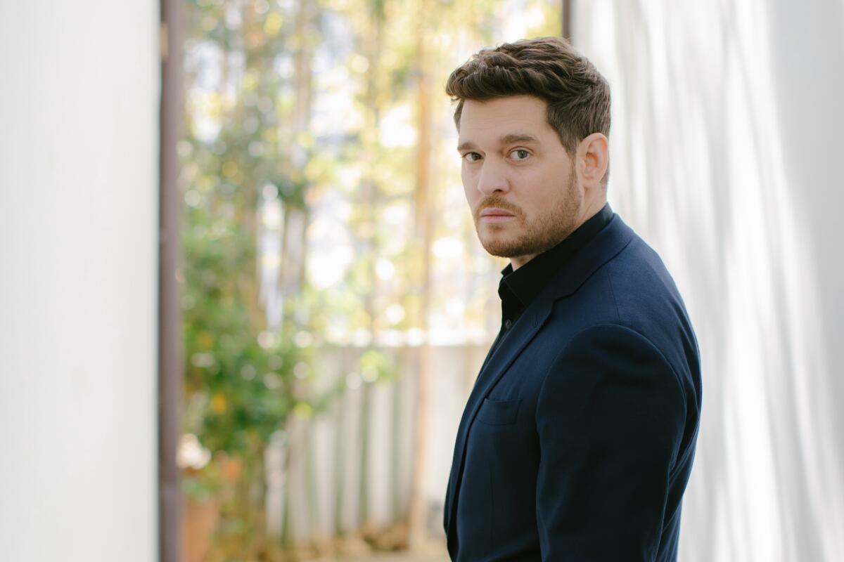 Michael Bublé in a dark blue suit stands next to a window with a blurred outdoor view and a white curtain on one side