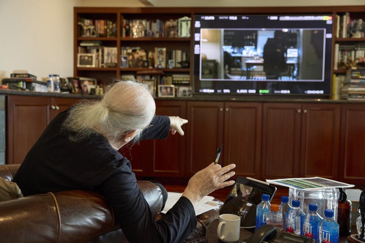John Carpenter, seen from the back, sits on a brown couch and his hand outstretched to his TV screen.