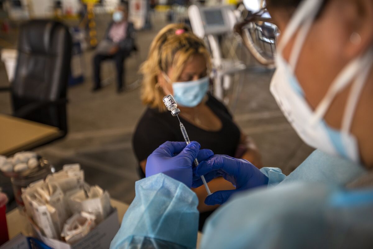 A young girl in a mask sits in a chair as a technician prepares an injection 