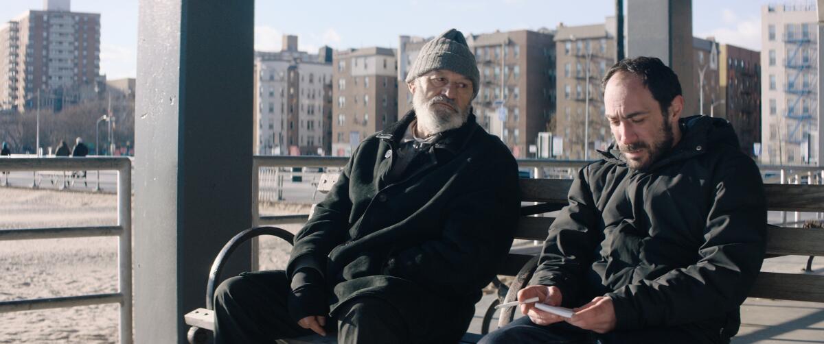 Two men sit on a bench at the beach with buildings behind them in the movie “Brighton 4th”